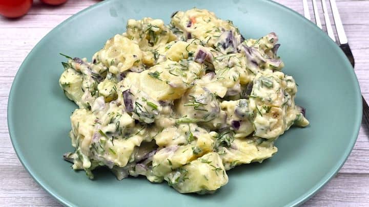 Plated creamy potato salad garnished with fresh herbs next to a cherry tomato.