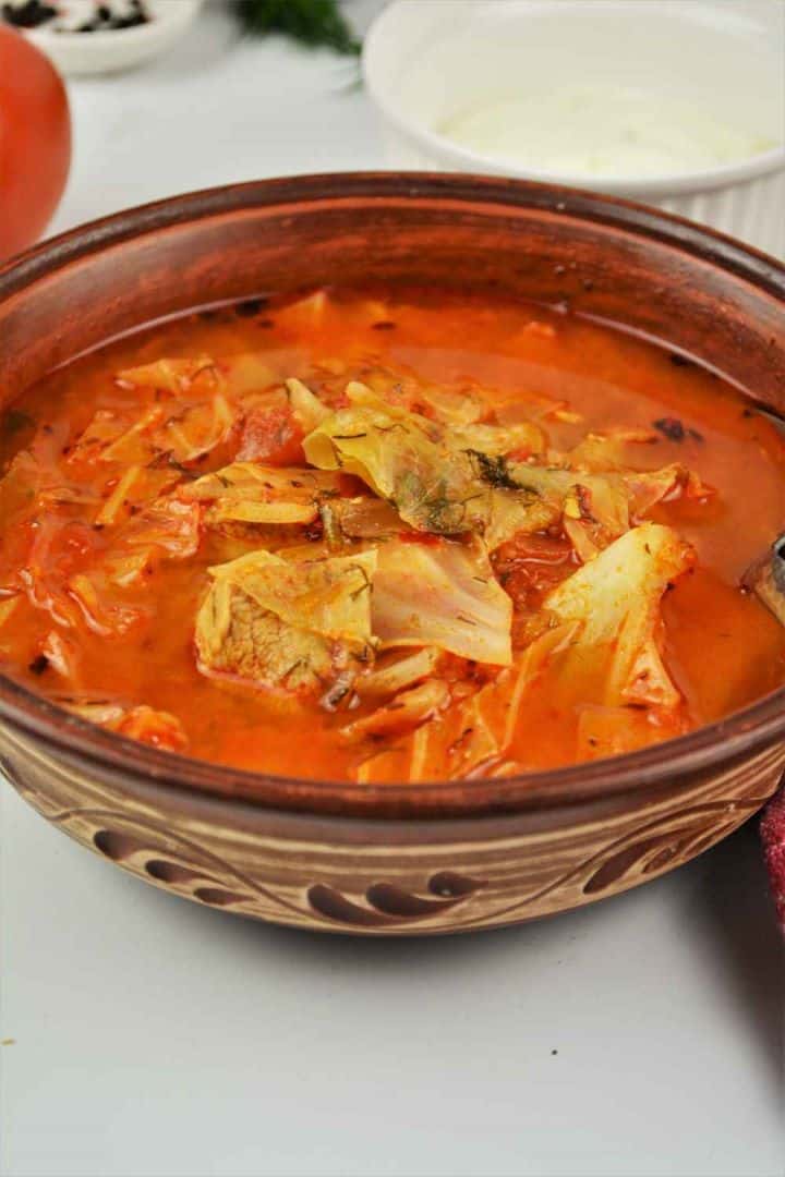 A rustic brown bowl filled with hot cabbage soup with pork meat, topped with fresh sour cream.