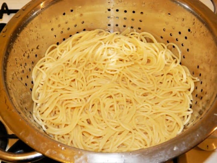 Draining and rinsing cooked spaghetti under cold running water in a metal colander.