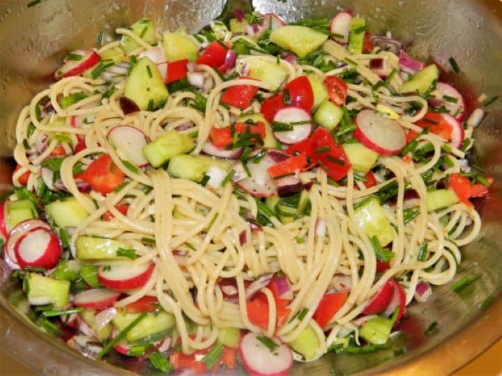Using kitchen tongs to gently toss warm spaghetti noodles with chopped vegetables and lemon-herb dressing.