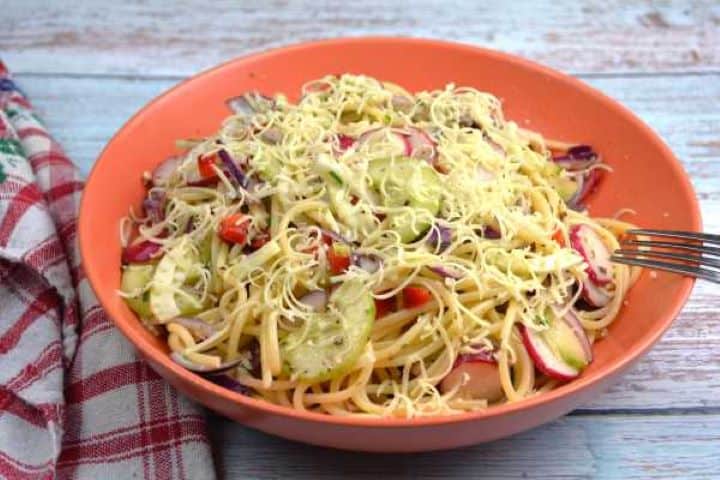 A refreshing orange bowl of vegetarian spaghetti salad mixed with crunchy vegetables and topped with shredded cheddar cheese and extra chives.