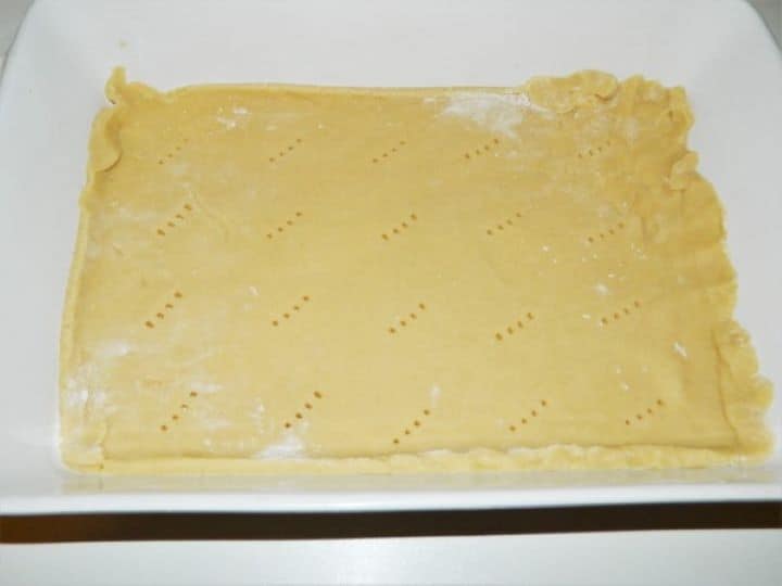 A photograph showing a rectangular baking pan lined with parchment paper, where the first rolled-out sheet of shortcrust pastry is being evenly pricked across the surface with a fork, demonstrating how to prevent air pockets before adding the apple filling.