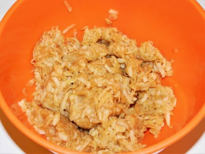 A close-up action shot showing a hand firmly squeezing moisture out of freshly grated apples into a ceramic bowl, illustrating the essential grandmother's technique to prevent a soggy bottom crust in a layered grated apple cake.