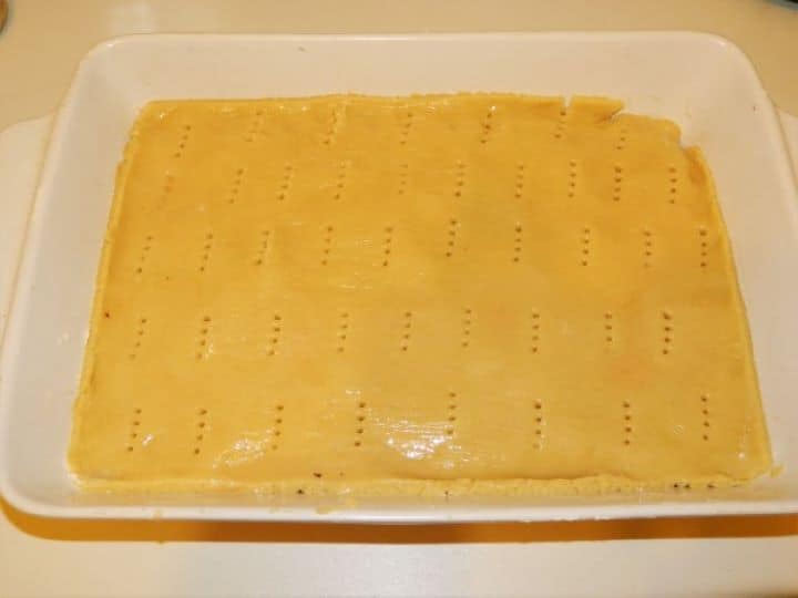 A view of the layered cake assembly in a baking pan. The thick, spiced grated apple filling is now covered by the second rolled-out dough sheet, which is being pricked with a fork and glazed with a beaten egg using a pastry brush, ready for the preheated oven.