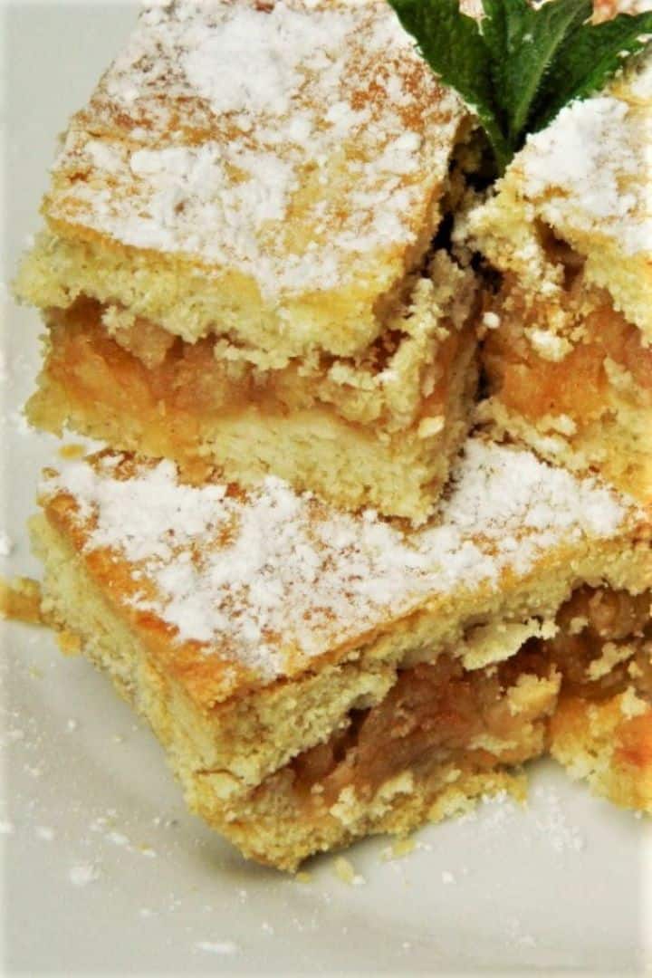 A close-up of a perfectly baked traditional Hungarian layered apple cake (Almás Pite) cut into neat squares, showing the golden-brown shortcrust pastry top dusted with powdered sugar and the dense, jam-like, spiced grated apple filling, ready to serve with coffee on a white plate.