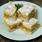 A close-up of a perfectly baked traditional Hungarian layered apple cake (Almás Pite) cut into neat squares, showing the golden-brown shortcrust pastry top dusted with powdered sugar and the dense, jam-like, spiced grated apple filling, ready to serve with coffee on a white plate.