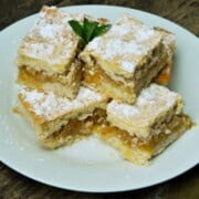 A close-up of a perfectly baked traditional Hungarian layered apple cake (Almás Pite) cut into neat squares, showing the golden-brown shortcrust pastry top dusted with powdered sugar and the dense, jam-like, spiced grated apple filling, ready to serve with coffee on a white plate.