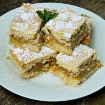 A close-up of a perfectly baked traditional Hungarian layered apple cake (Almás Pite) cut into neat squares, showing the golden-brown shortcrust pastry top dusted with powdered sugar and the dense, jam-like, spiced grated apple filling, ready to serve with coffee on a white plate.