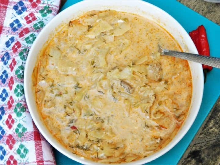 A deep rustic bowl filled with thick Lucskos Kaposzta cabbage soup, served with a side of hot chili peppers and fresh bread.
