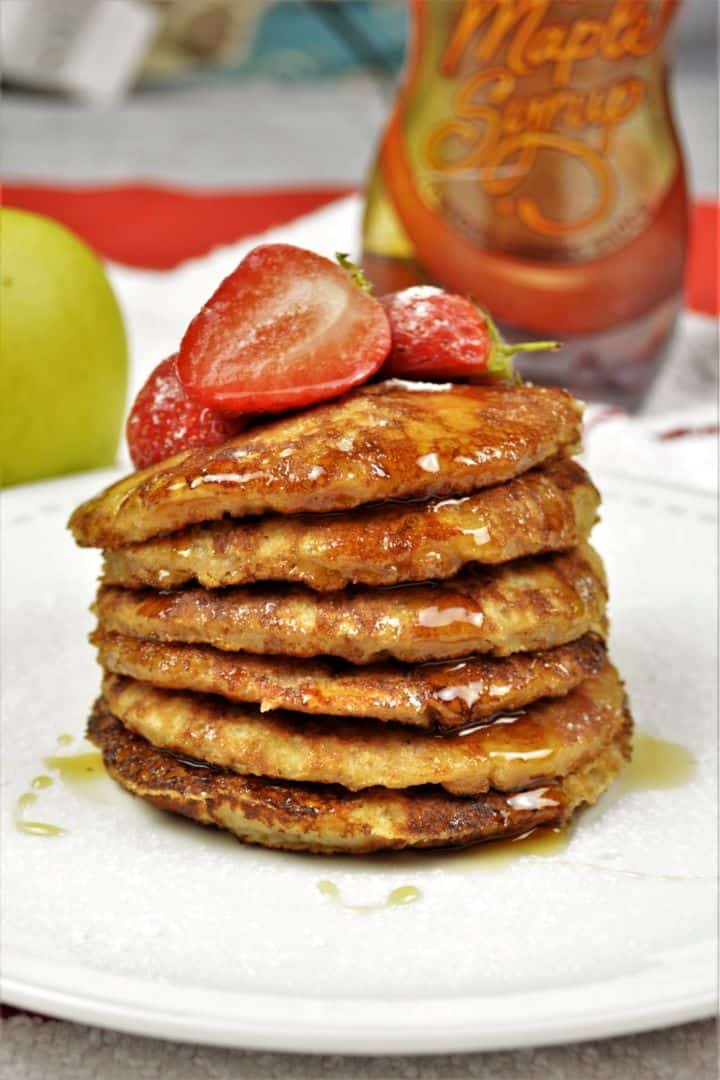 Easy Cinnamon Apple Pancakes Recipe-Served on Plate With Maple Syrup and Strawberry A tall stack of fluffy cinnamon apple pancakes on a white plate, lightly dusted with powdered sugar and drizzled with maple syrup, showing raisins mixed into the dough.
