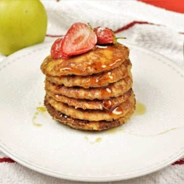 A tall stack of fluffy cinnamon apple pancakes on a white plate, lightly dusted with powdered sugar and drizzled with maple syrup, showing raisins mixed into the dough and strawberries on top.
