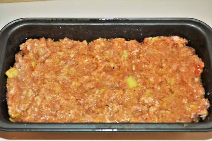 Pressing the first layer of seasoned pork mince evenly into the bottom of a greased loaf baking pan.