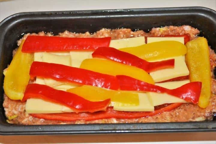 Layering sliced red kapia peppers, yellow bell peppers, and thick cheddar cheese strips down the center of a pork meatloaf base.