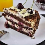 Slice of easy Black Forest cake on a white plate showing layers of chocolate sponge, whipped cream, and cherries.