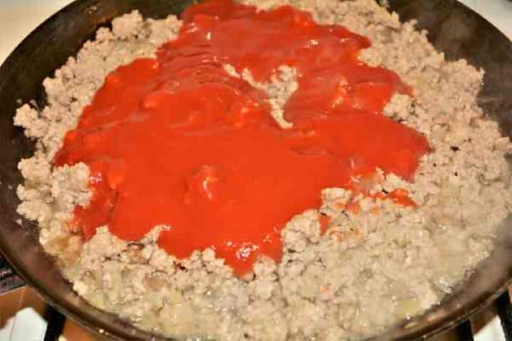 Easy Potato Moussaka Recipe-Pouring Tomato Passata Over Fried Pork Mince in the Pan Pouring tomato passata and dried herbs over browned minced pork in the pan.