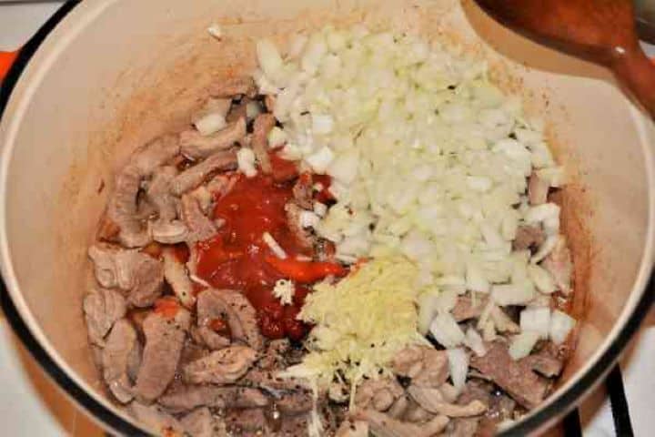 Brasov Roast Recipe-Simmering Pork Stew in the Pot Stirring chopped onions, minced garlic, tomato passata, and red goulash paste into the browned pork shoulder.