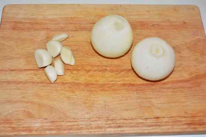 Brasov Roast Recipe-Cleaned and Peeled Onions and Garlic Cloves Two peeled yellow onions and six garlic cloves prepared on a wooden chopping board.