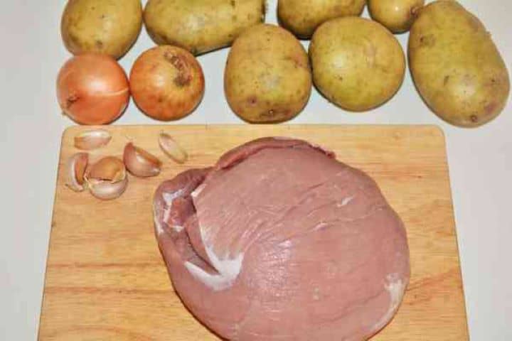 Brasov Roast Recipe-Pork Shoulder, Potatoes, Onions and Garlic Cloves Raw diced pork shoulder, chopped onions, minced garlic, and cubed potatoes prepped on a wooden cutting board.