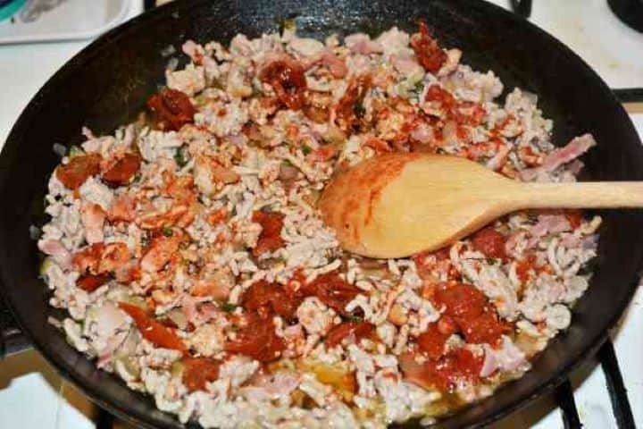 Stuffed Aubergines Recipe-Adding Sundried Tomatoes on the Simmering Mince in the Pan Minced pork, crispy smoked bacon pieces, and diced onions simmering with sun-dried tomatoes and thyme in a large frying pan.