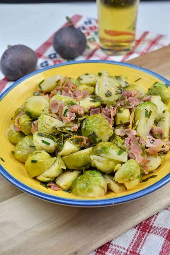 Brussels Sprouts With Lemon-Served in the Bowl on the Chopping Board With a Glass of Beer Easy Brussels sprouts with lemon, smoked bacon, and chopped chives served in a yellow bowl on a wooden board.