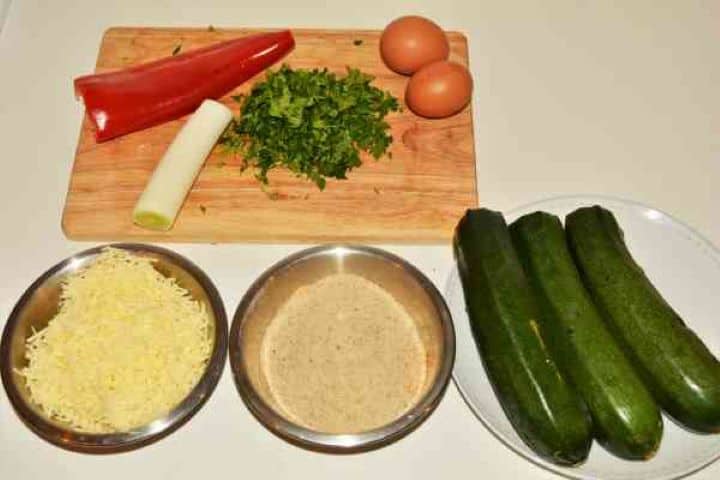 Zucchini Balls Recipe-Zucchini, Breadcrumbs, Grated Cheddar, Eggs, Half Kapia Pepper, Leek and Parsley Ingredients for making fried zucchini balls on a table including courgettes, grated cheddar cheese, breadcrumbs, eggs, leek, and half a red kapia pepper.