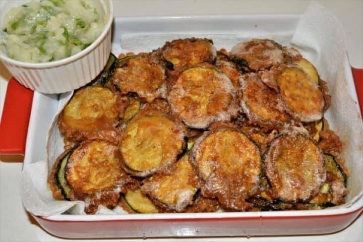 Battered Fried Zucchini-Fried Zucchini Slices in the Baking Dish With Garlic Cream in the Bowl Fried beer battered zucchini rounds resting on paper towels next to a bowl of garlic cream.
