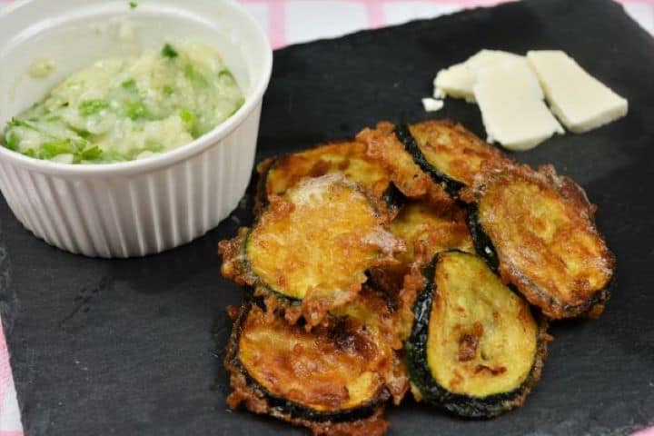 Battered Fried Zucchini-Served on Slab With Garlic Cream and Cheddar Cheese Warm beer battered zucchini rounds arranged on a plate with homemade garlic dip and cheddar slices.