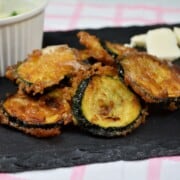 Golden beer battered fried zucchini slices served on a plate with creamy garlic dip and cheddar cheese.