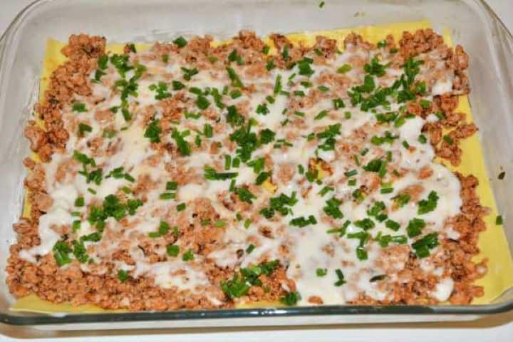 Turkey Lasagna With White Sauce-Cheese Sauce and Chopped Chives on the First Filling Layer in the Baking Tray Pouring white cheese sauce and scattering chopped chives over the seasoned turkey meat.