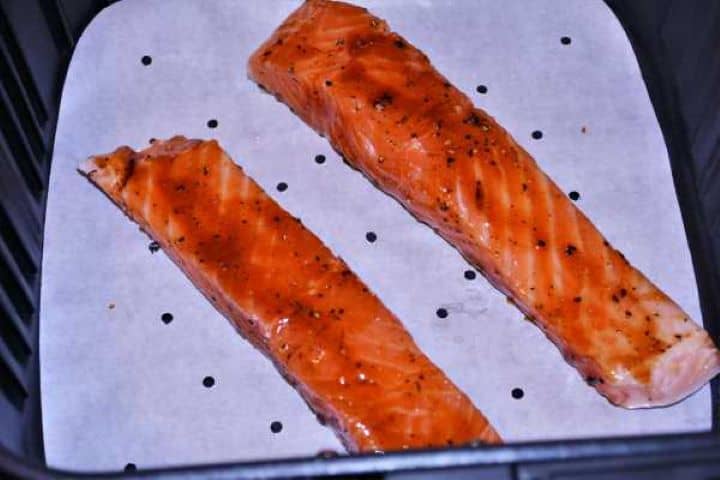 Salmon Fillets in the Air Fryer Two glazed salmon fillets placed skin-side down on a piece of perforated white parchment paper inside a clean air fryer basket.