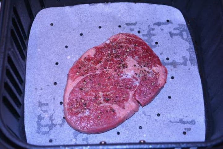 Ribeye Steak in Air Fryer-Seasoned Ribeye Steak in the Air Fryer Basket Seasoned ribeye steak placed on parchment paper inside the hot air fryer basket before cooking.