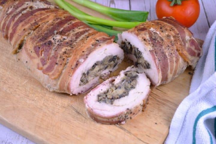 Stuffed Turkey Tenderloin Recipe-Sliced and Served on the Chopping Board Sliced bacon wrapped stuffed turkey tenderloin arranged on a wooden chopping board garnished with fresh red tomatoes and sliced spring onions.