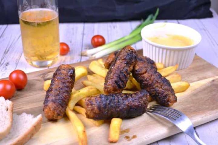 Tender grilled Romanian mititei (skinless sausages) served on a wooden platter alongside crispy fries and smooth yellow mustard.