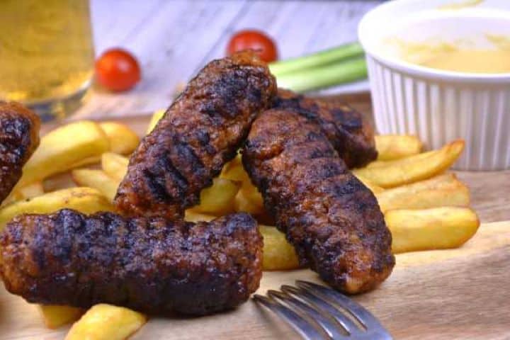 Served Freshly cooked homemade skinless sausages arranged on a rustic wooden cutting board with golden French fries and a side of yellow mustard.