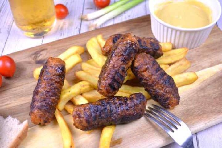 Serving Platter of grilled Romanian skinless sausages with crusty bread slices, yellow mustard, fries, and cold beer.
