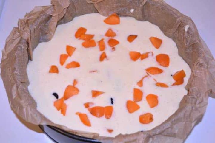 Prepared Pouring smooth flourless yoghurt cake batter into a parchment-lined baking tin and scattering sweet raisins and sliced apricots on top.