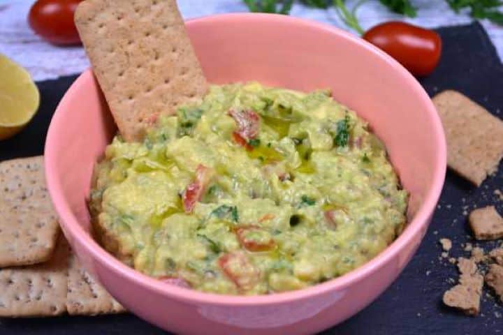 Served Overhead view of homemade guacamole without cilantro surrounded by crackers on a table.