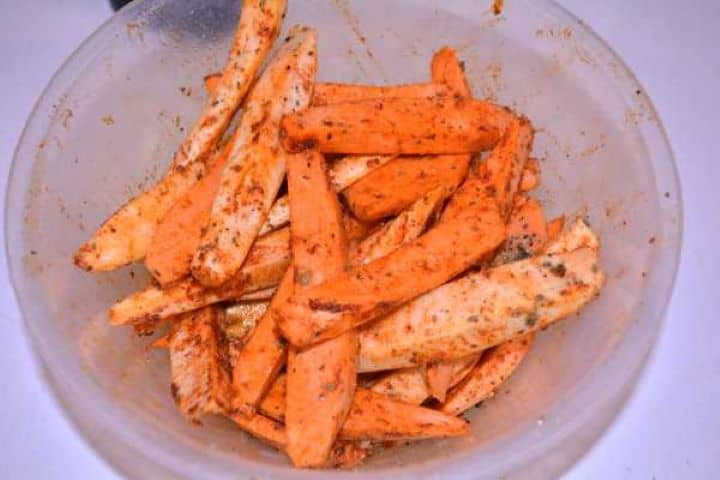 Seasoned Raw peeled sweet potato wedges tossed with olive oil, oregano, onion powder, and Hungarian paprika in a plastic mixing bowl.