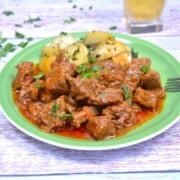 Authentic Hungarian beef paprikash with tender beef cubes and rich red sauce on a green plate with potatoes.