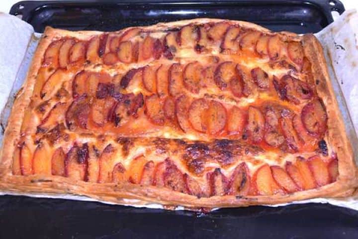 Ready Golden brown baked puff pastry plum tart with bubbling fruit juices fresh from the oven on a baking tray.