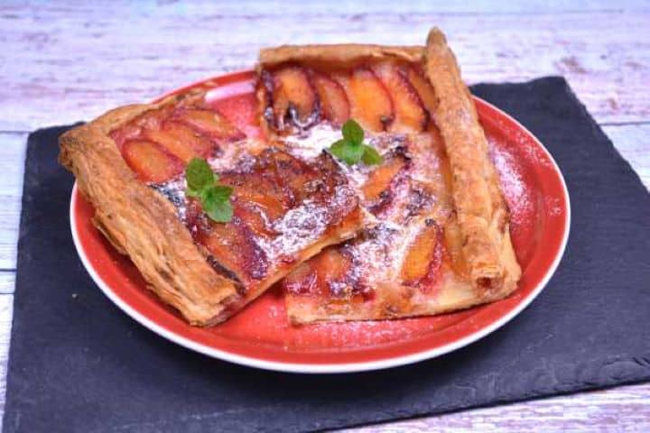 Serve Warm slices of puff pastry plum tart dusted with powdered sugar and served on a red ceramic plate.