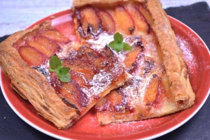 Served Close-up view of a slice of warm puff pastry plum tart showing flaky buttery pastry layers and caramelized plums dusted with powdered sugar.