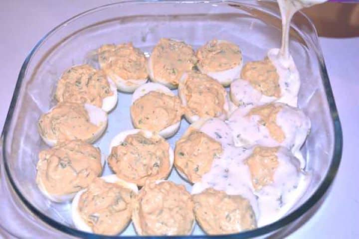 Pouring Pouring creamy homemade yogurt mayonnaise sauce over stuffed deviled eggs in a glass dish.