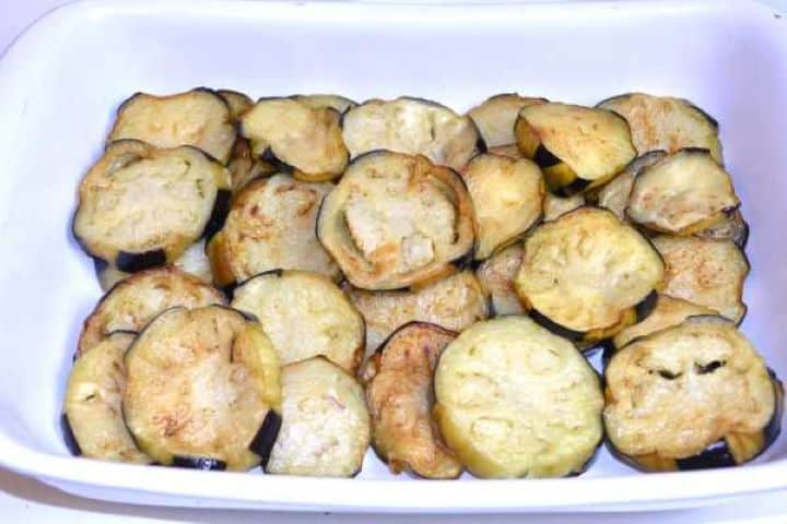 First Arranging a single layer of fried golden aubergine slices in a ceramic baking dish.