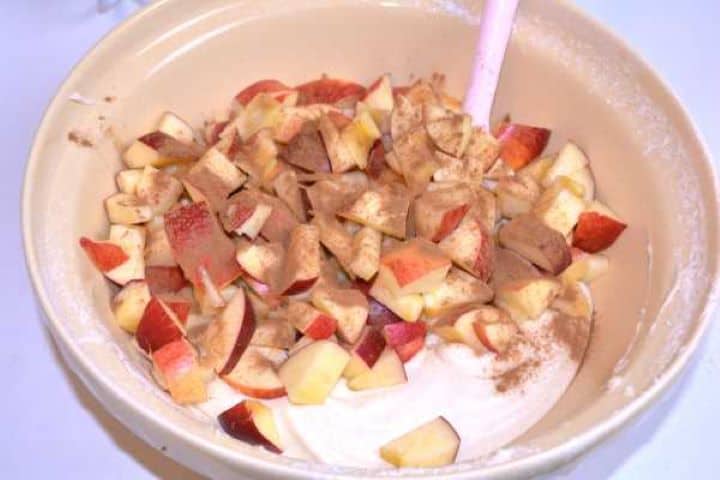Diced unpeeled apples and ground cinnamon sprinkled over the cake batter in a bowl.