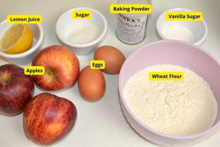 Ingredients for easy grated apple fritters including fresh apples, flour, eggs, sugar, baking powder, and lemon on a counter.