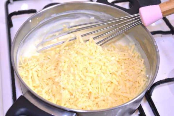 cheddar Adding freshly grated cheddar cheese into the thickened warm milk mixture in a pot.