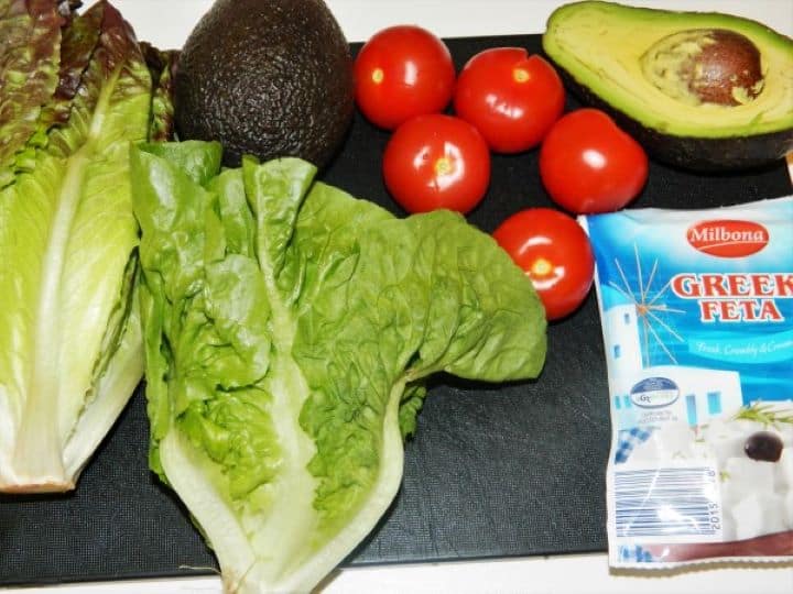 Fresh salad ingredients including romaine lettuce, ripe Hass avocado, cherry tomatoes, and Greek feta cheese block.