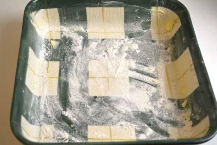 Greasing a 24 by 24 cm square ceramic baking dish with butter and dusting with flour for a rustic cake crust.