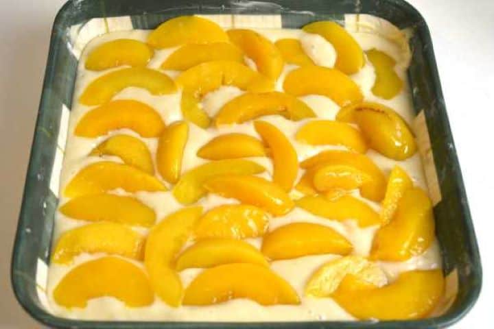 Arranging canned peach slices in a pattern on top of thick yogurt sponge cake batter in a square dish.